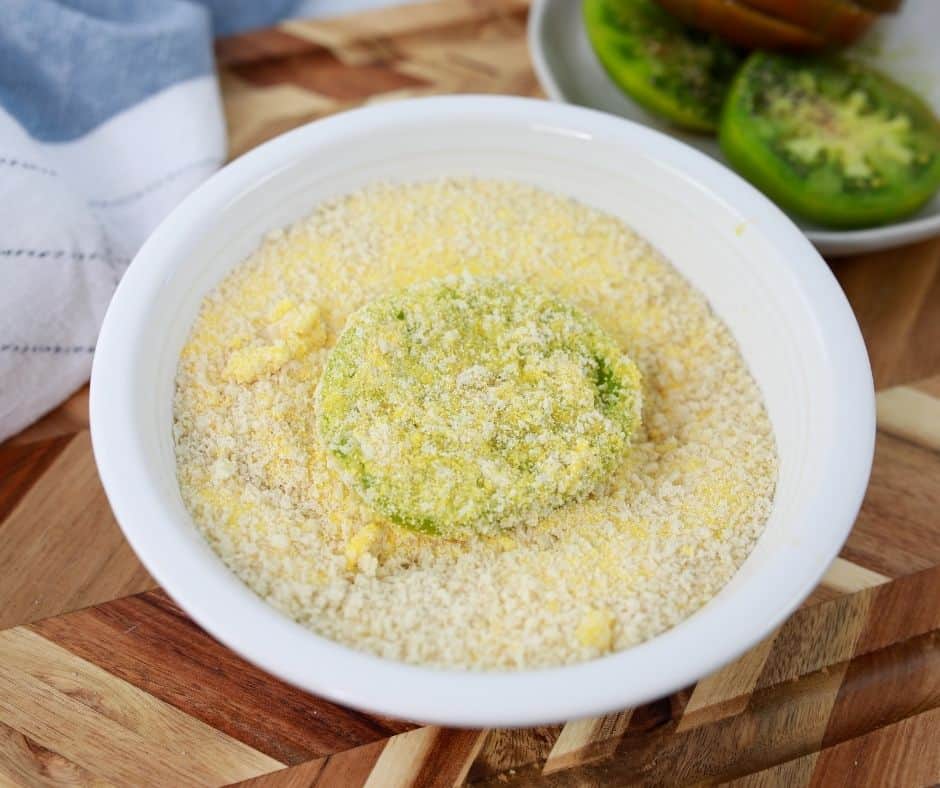 A white bowl filled with panko breadcrumbs and cornmeal with a green tomato slice submerged in it. 