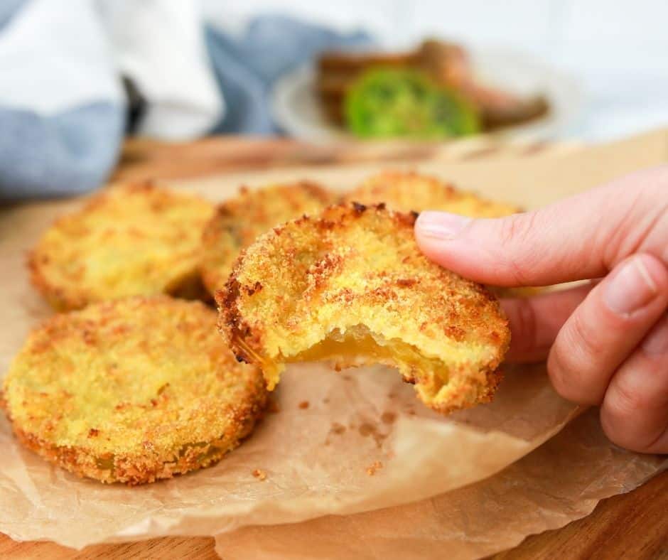 Close up of a hand holding an air fryer fried breaded green tomato, with more fried green tomatoes in the background on greaseproof paper on a chopping board. 