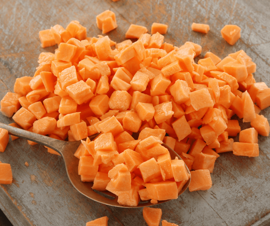 Carrots are diced into small cubes and placed onto a wooden chopping board. There is a large silver serving spoon underneath the pile with some of the carrots on top of it.