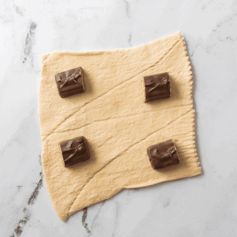Hands wrapping biscuit dough around Snickers pieces, sealing the edges, with an air fryer preheating in the background.