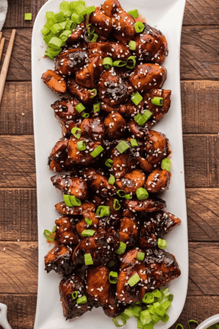 Golden, crispy air fryer firecracker chicken bites coated in sweet and spicy sauce, garnished with sesame seeds and green onions.