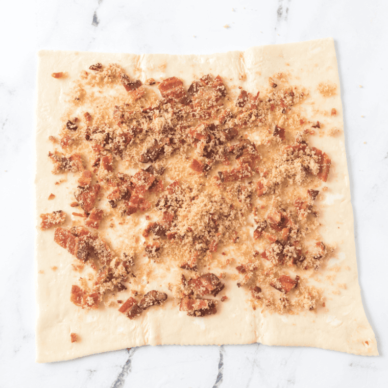 Spreading softened butter over puff pastry and topping with brown sugar, chili flakes, crispy bacon, and shredded cheddar cheese for breakfast pinwheels