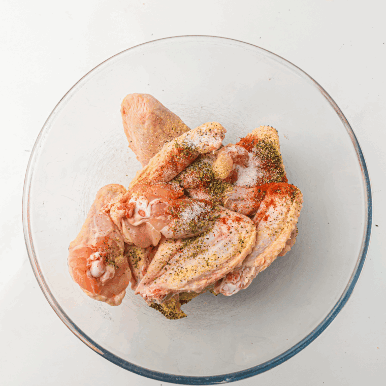 Chicken wings being tossed with olive oil in a large bowl, while a small bowl holds a mix of salt, black pepper, garlic powder, onion powder, paprika, and cayenne pepper seasoning
