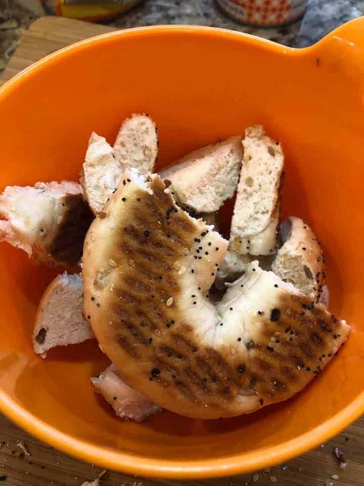 An orange bowl is filled with chunks of broken up seed coated bagels.