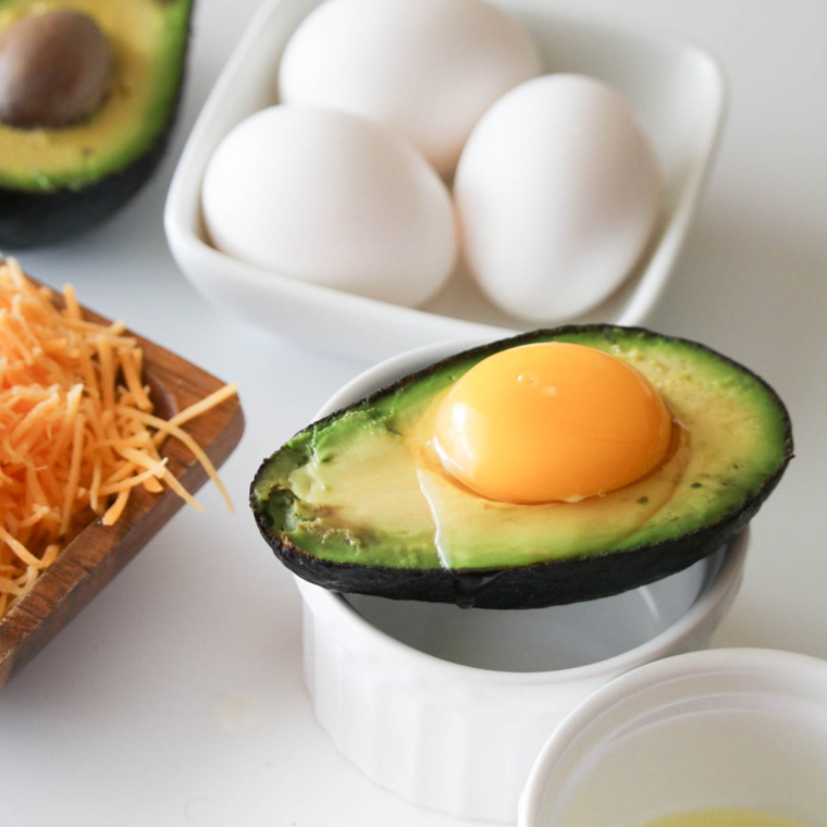 closeup: a raw egg cracked into an avocado half that is sitting on a white ramekin