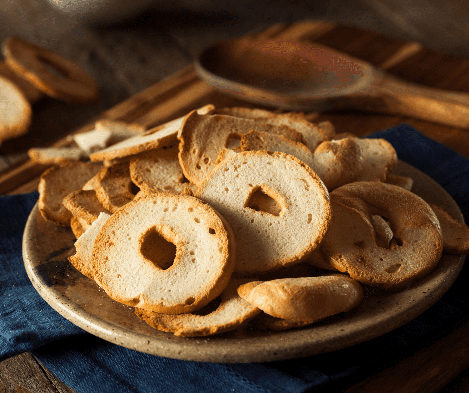 A ceramic plate is filled with bagel chips slices length ways so they are thins full, golden brown circles. There is a wooden spoon in the background and some extra bagel chips.