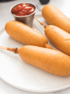 four cooked corn dogs with ketchup on a white plate