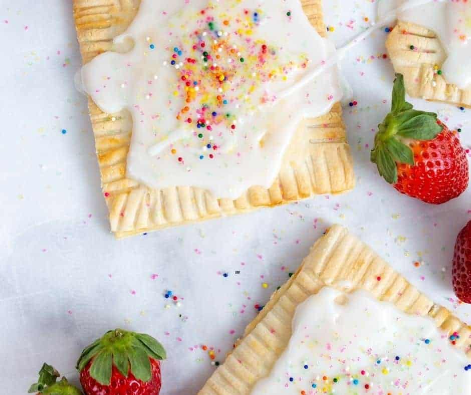 2 partially seen Air Fryer Pop-Tarts sitting on a white counter. Both pop tarts have white icing and sprinkle on top. There are some leftover strawberries on the counter also.