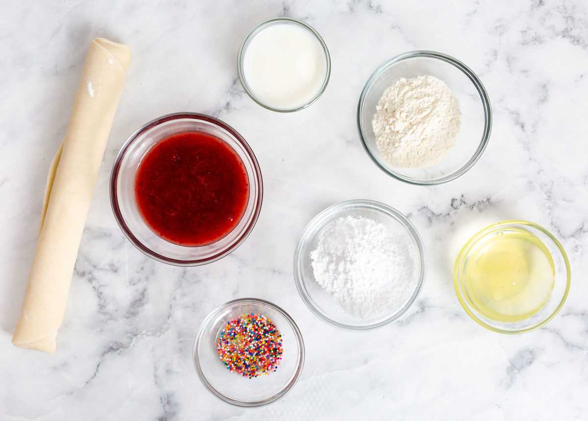 Ingredients Needed For homemade Pop-Tarts, each ingredient is in it's own glass bowl, sitting on a white marble counter. There is flour, sugar, egg whites, strawberry jam, sprinkles and a rolled up piece of pastry.