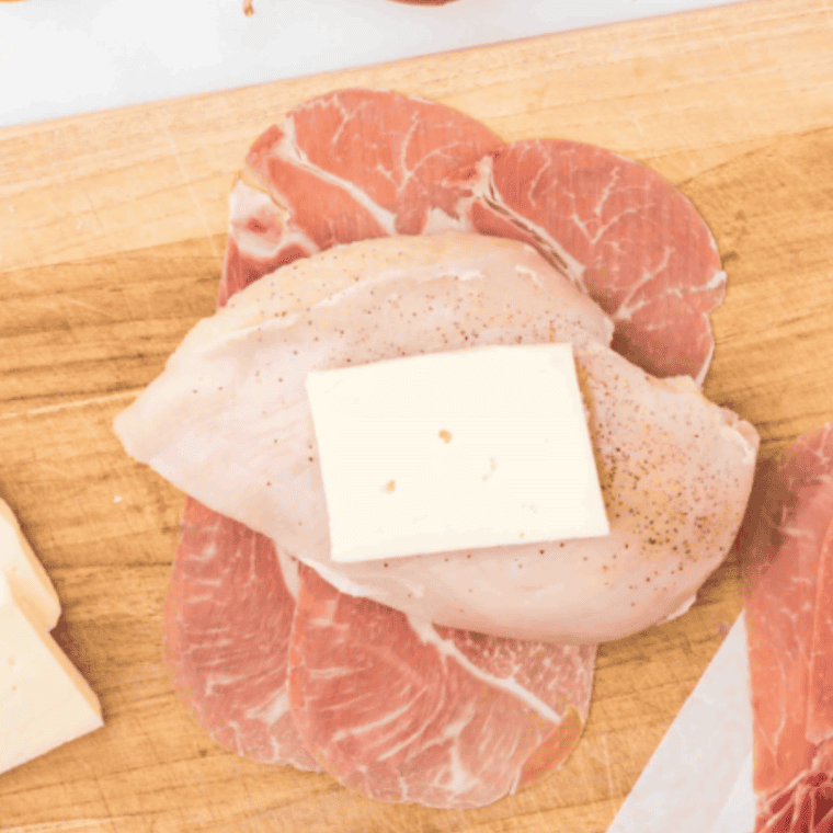 Chicken breast topped with fontina cheese and sage leaf, wrapped in overlapping prosciutto slices on a prep surface.