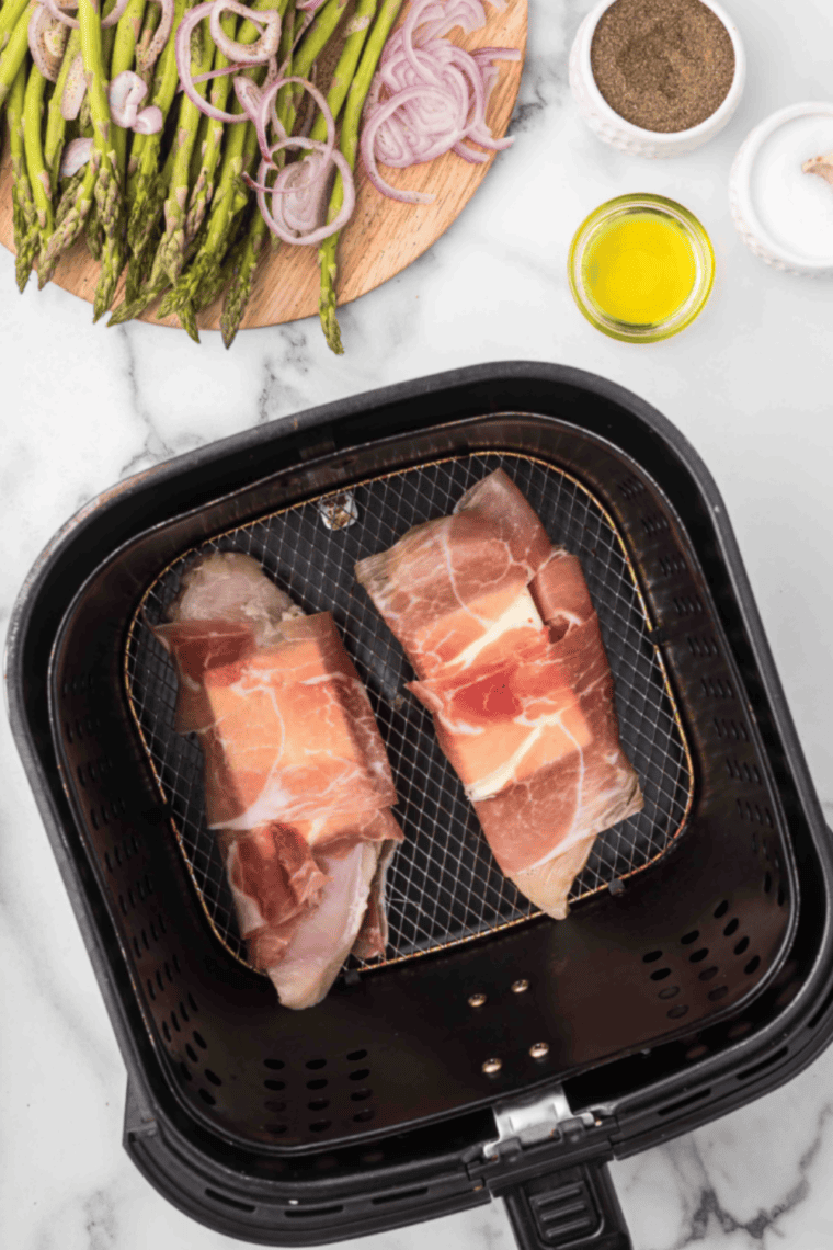 Prosciutto-wrapped chicken placed in air fryer basket with asparagus and shallots surrounding it, ready to cook