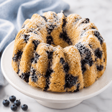 Copycat Nothing Bundt Cakes lemon blueberry bundt cake with piped cream cheese frosting ribbons, one slice cut to show blueberries in the crumb