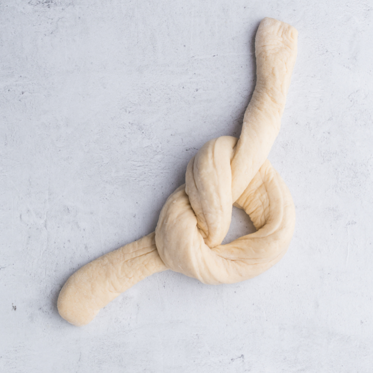 Dough ropes being tied into loose overhand knots and tucked underneath, forming classic garlic knot shapes.