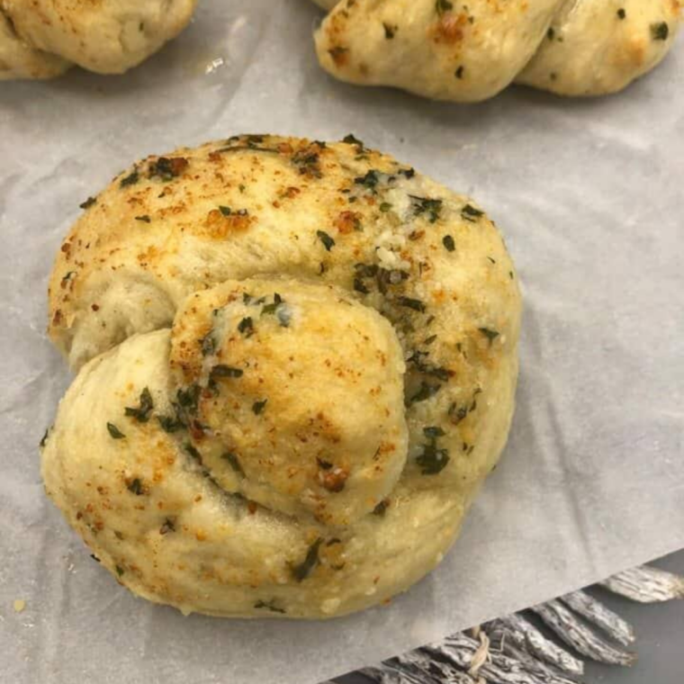 Garlic knots being tossed in a bowl of melted garlic butter with herbs and cheese.