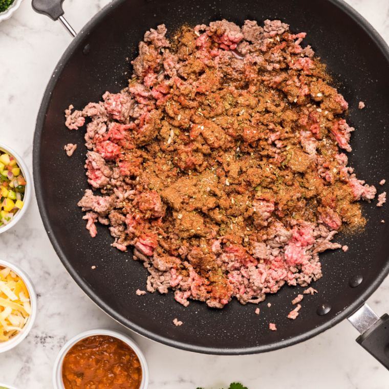 Homemade taco seasoning sprinkled over ground beef browning in a skillet for air fryer taco cups