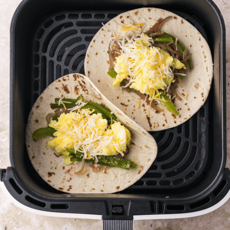 Assembling breakfast tacos by adding sautéed vegetables, scrambled eggs, and shredded cheese to tortillas in the air fryer basket.