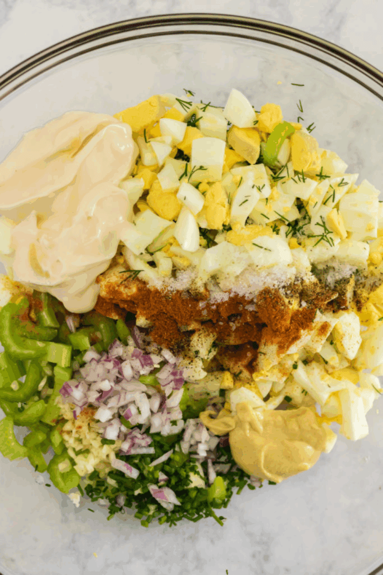 Mayonnaise, garlic, lemon juice, mustard, and seasonings being mixed in a small bowl, then combined with chopped eggs and veggies for egg salad, ready to serve on toasted bread or in a chilled dish.