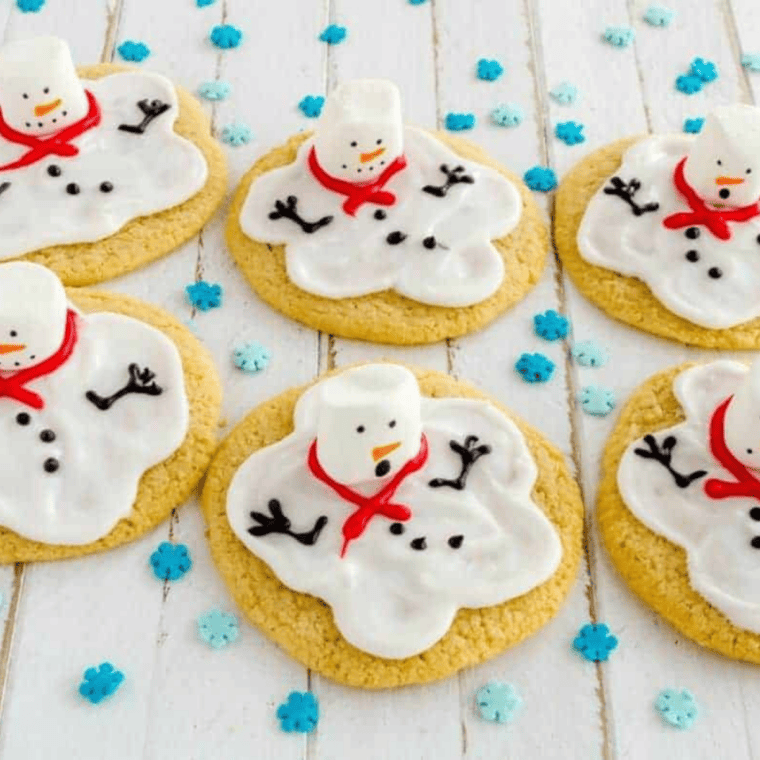 A row of whimsical melting snowman cookies with white icing puddles, marshmallow heads, and tiny orange carrot noses sitting on a cooling rack.