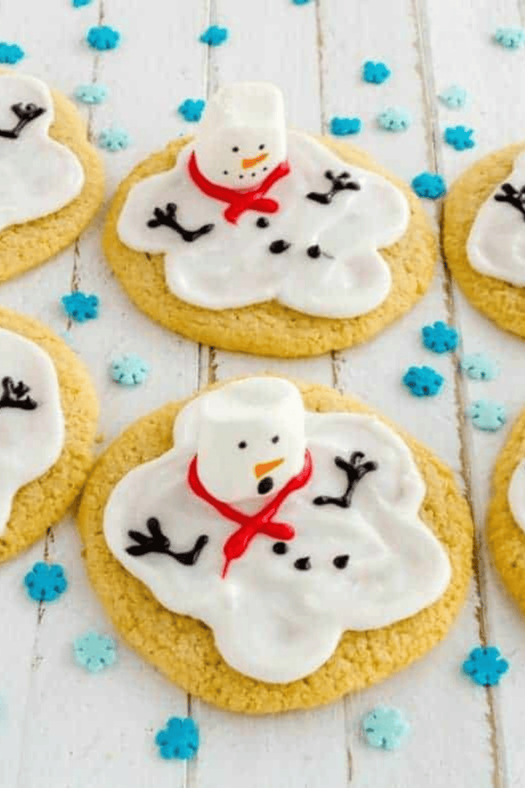 Adorable melting snowman cookies on a cooling rack, featuring white icing "puddles," marshmallow heads with coal eyes, and tiny orange carrot noses.