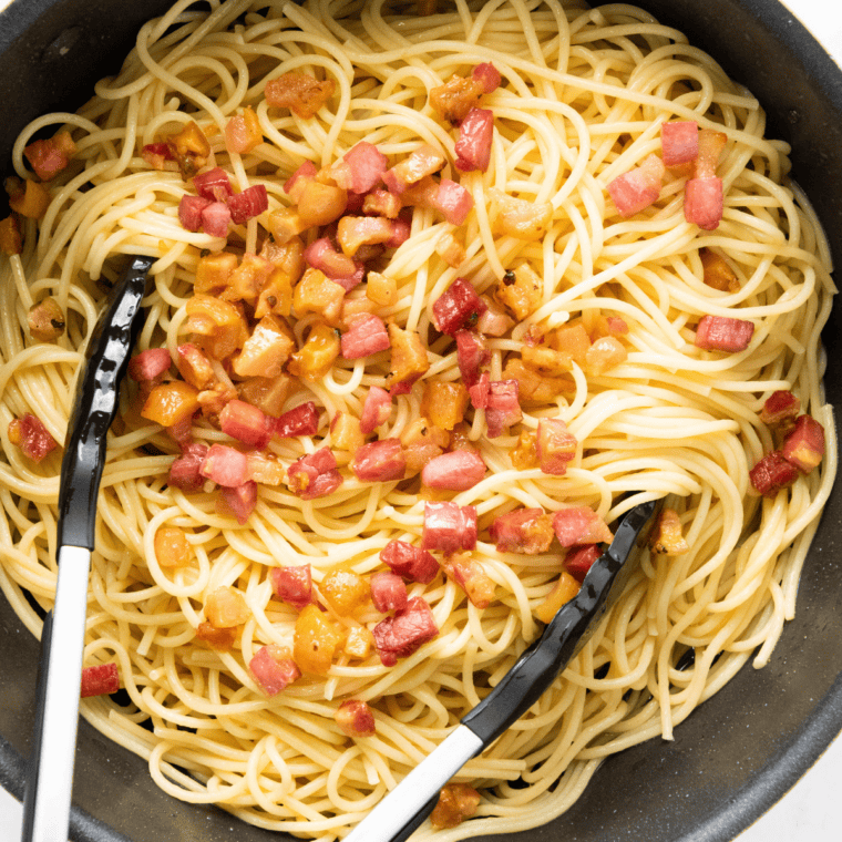 Egg and cheese mixture being tossed with hot spaghetti using tongs to create silky carbonara sauce