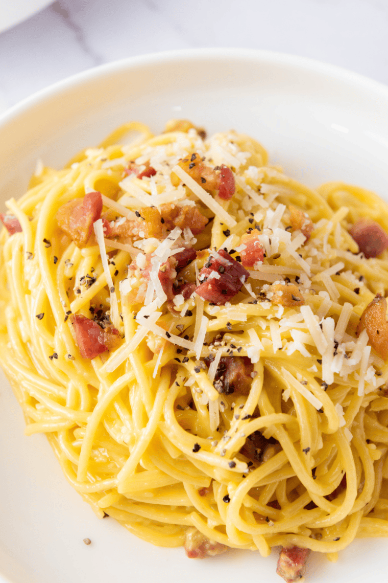 Bowl of creamy pasta carbonara with crispy bacon bits and cracked black pepper on top.