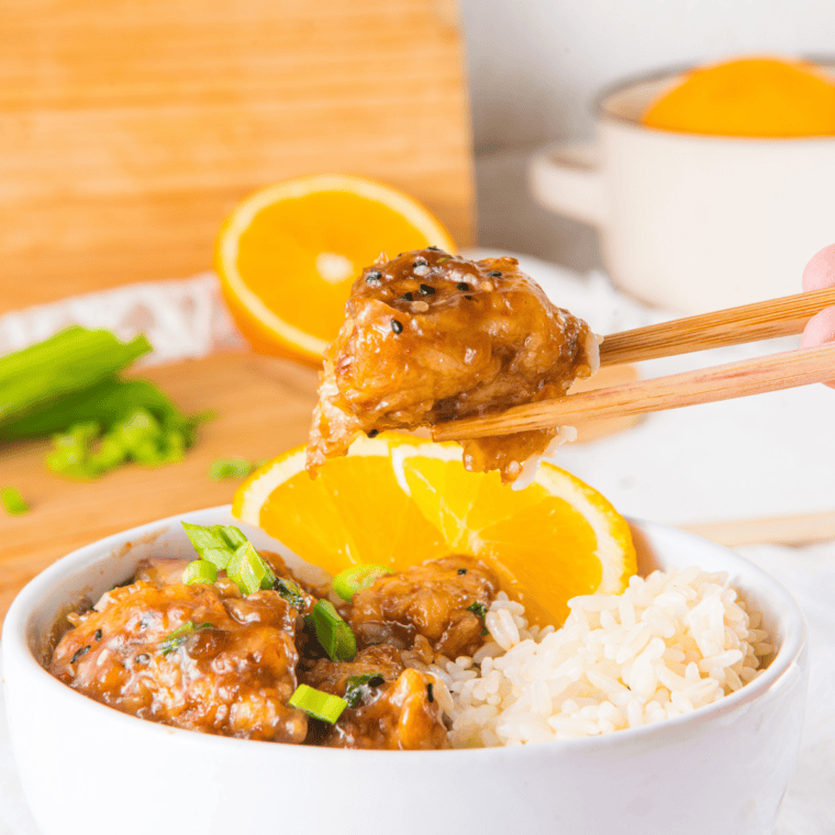 Slow cooker orange chicken over steamed white rice garnished with sesame seeds and green onions.