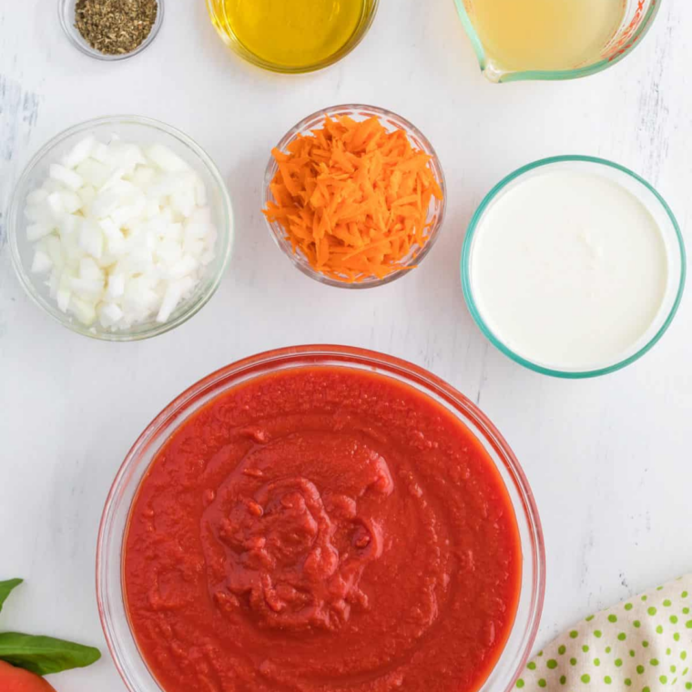Olive oil, diced onion, shredded carrots, dried basil, canned tomatoes, broth and heavy cream in bowls on a counter