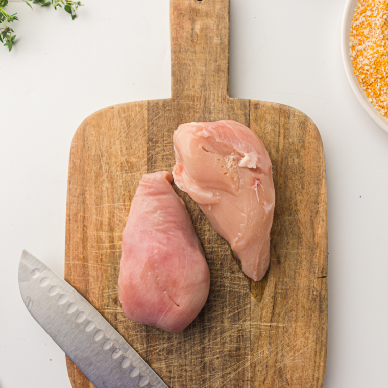 Chicken breast being sliced horizontally and pounded evenly between plastic wrap to create thin cutlets for air fryer breaded chicken.