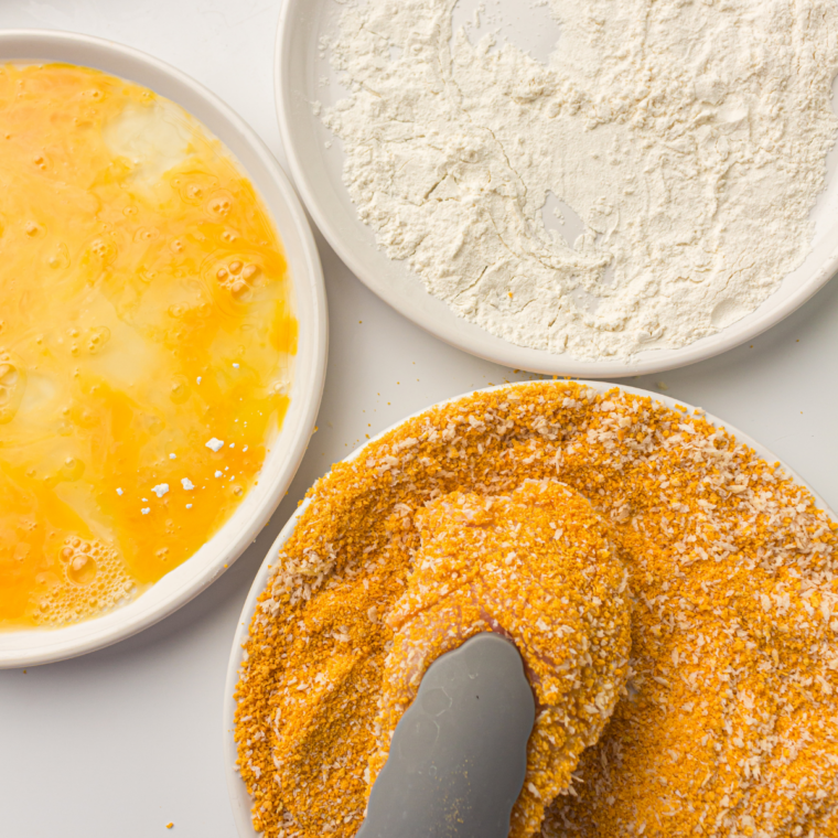 Chicken cutlet being coated in flour, dipped in egg, and pressed into seasoned panko breadcrumbs for a crispy breaded crust.