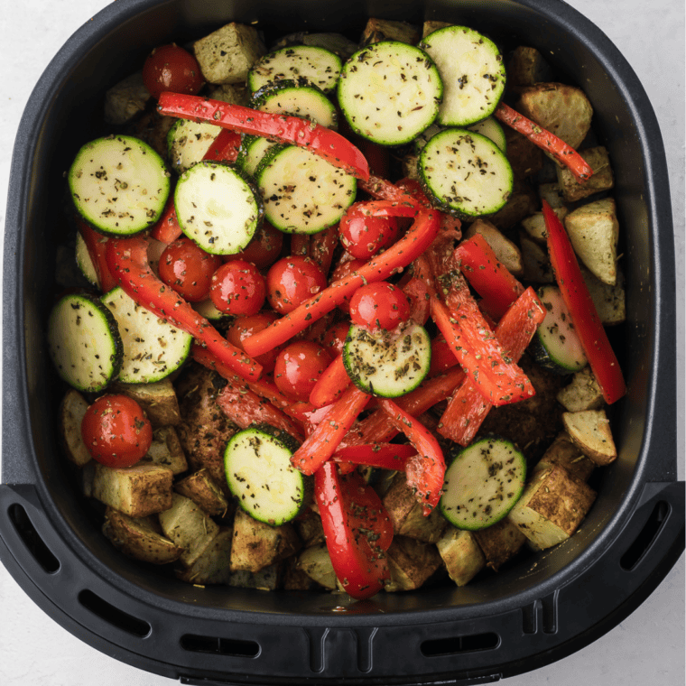 Adding seasoned vegetables to the air fryer basket with chicken and potatoes, cooking until tender and slightly charred, then serving hot with fresh herb garnish.