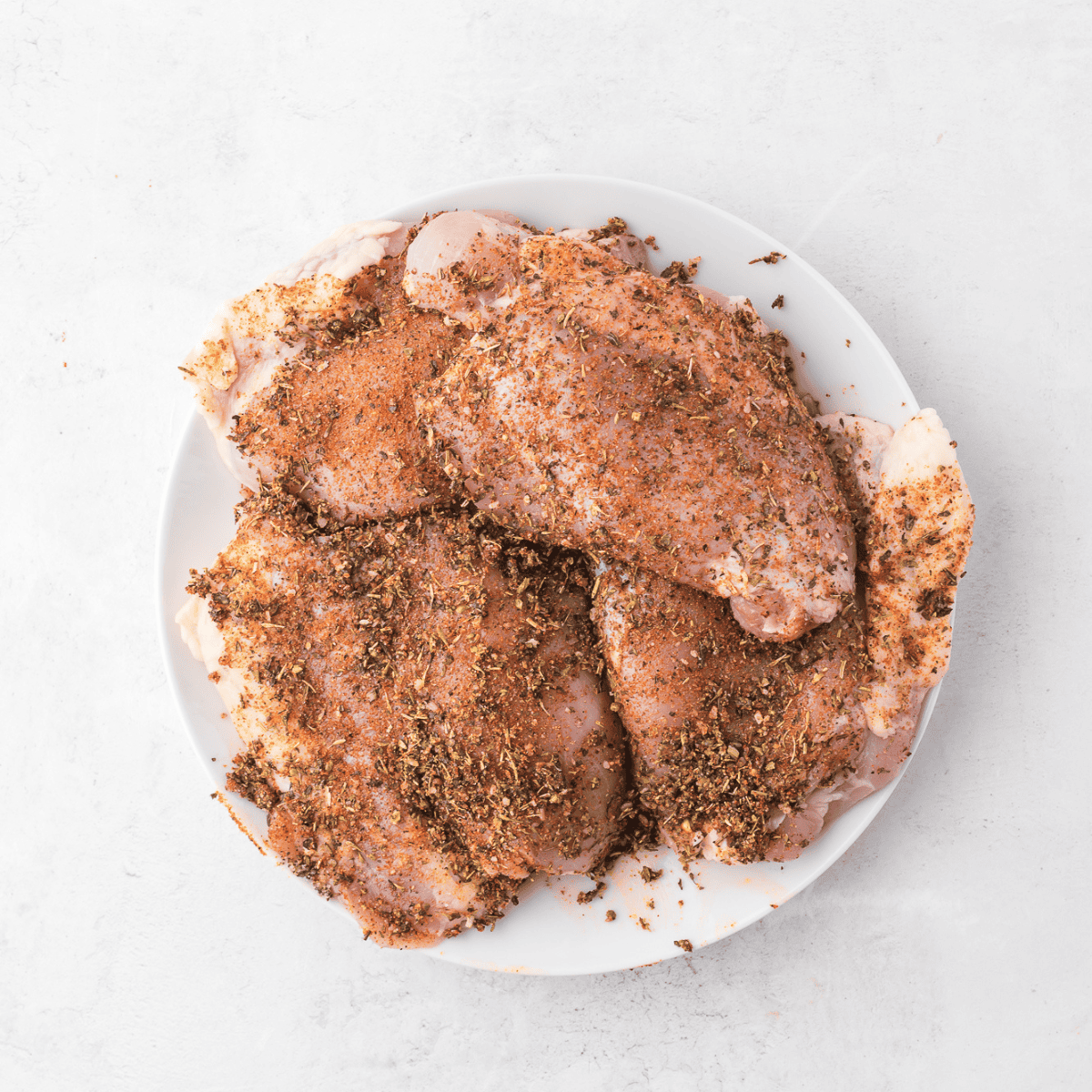 Mixing olive oil and Tuscan spices in a bowl, then rubbing the seasoning blend onto raw chicken thighs for even coating.