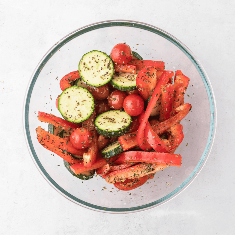 Tossing sliced bell pepper, zucchini, and cherry tomatoes with olive oil and dried herbs in a mixing bowl.