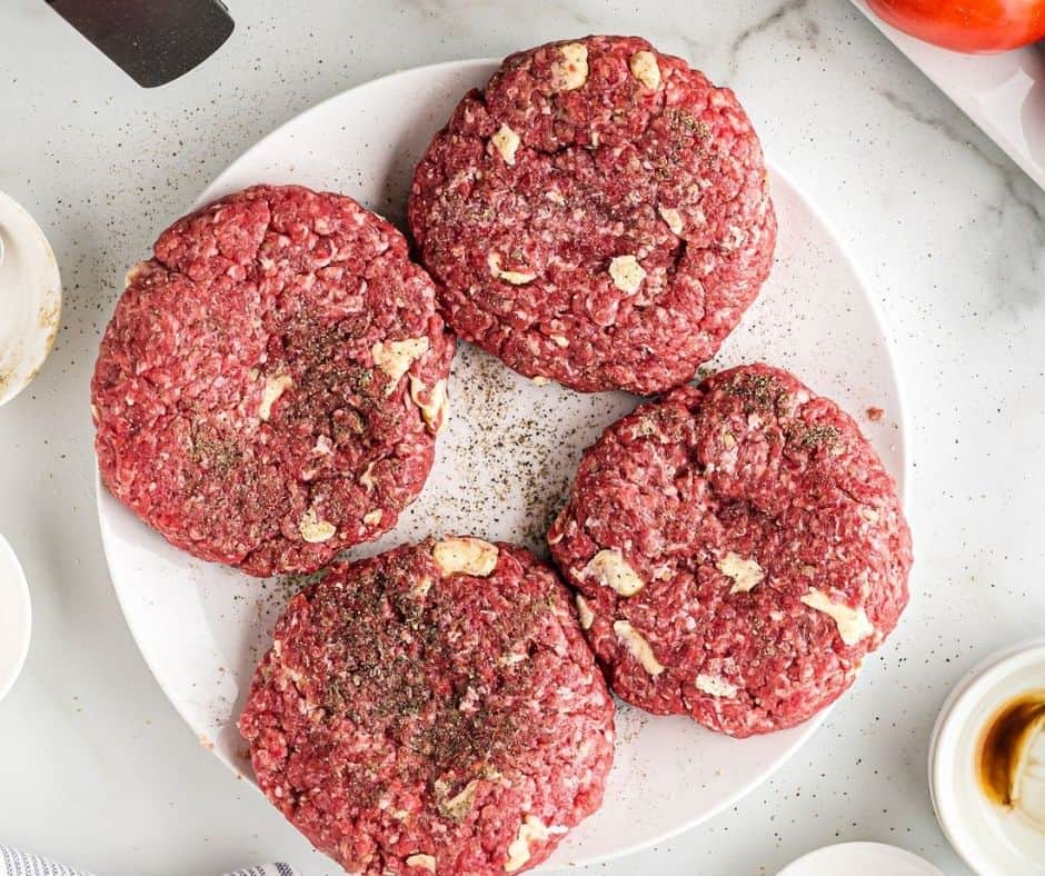 Top view of four raw burger patties on a white plate with salt and pepper sprinkled over them. 