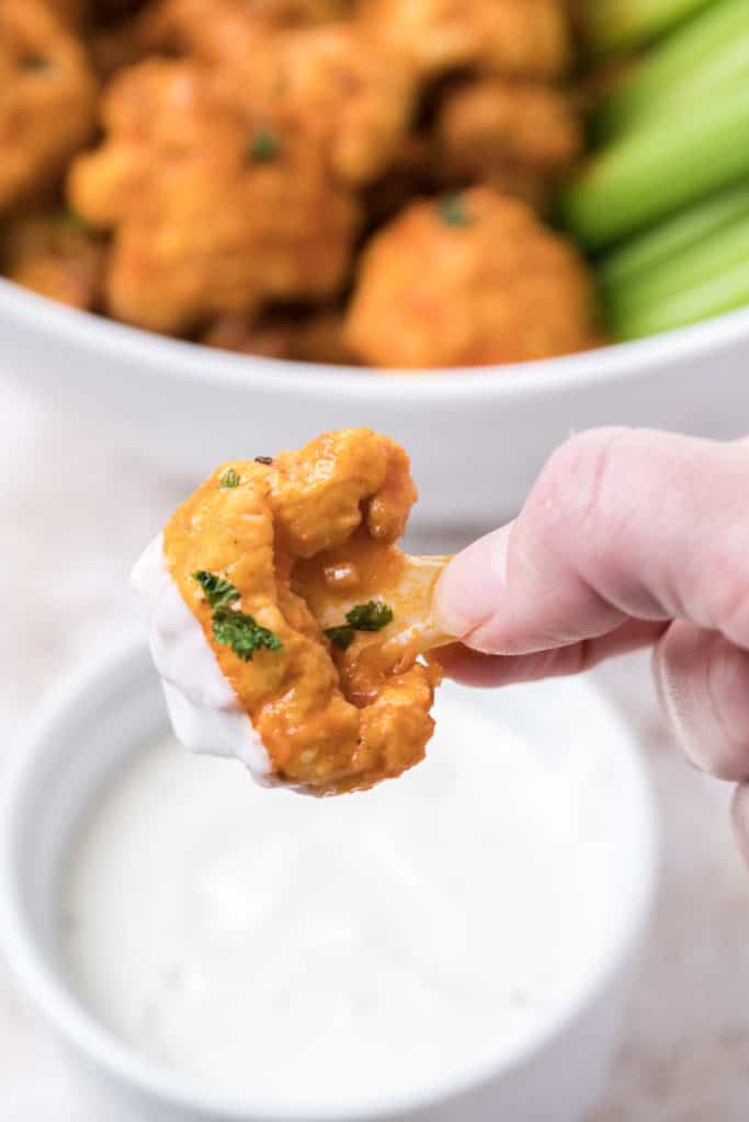 Buffalo Cauliflower Dipped in Blue Cheese Dressing. There is a white ramekin dish containing more blue cheese dressing and a large bowl in the background containing lots more air fryer buffalo cauliflower bites and celery sticks.