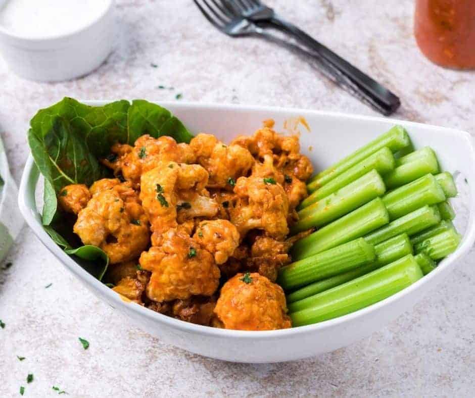 Air Fryer Buffalo Cauliflower Bites garnished with fresh herbs on large salad leave in a deep white bowl alongside a handful of celery sticks.