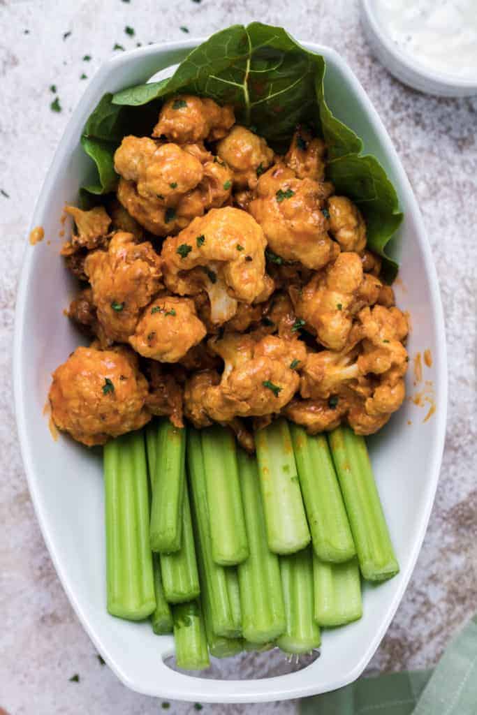 Air Fryer Buffalo Cauliflower Bites garnished with fresh herbs in a deep white bowl with salad leaves and celery sticks. There is a small bowl of blue cheese dressing (partial view) in the top right corner.