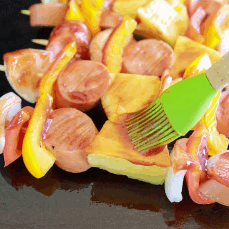  Grilled kielbasa kabobs sizzling on a hot griddle with BBQ sauce being brushed on.