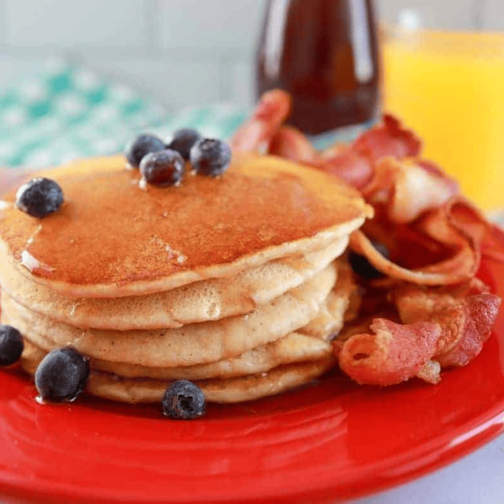 Blackstone Pancakes topped with syrup and blueberries on a red plate with bacon. 