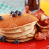 tack of golden pancakes with butter melting on top, dripping with maple syrup, blueberries scattered around, served on the red plate. ]