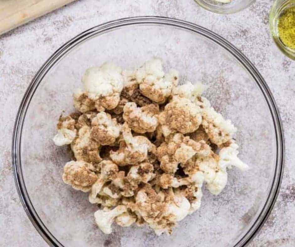 A large glass bowl sitting on a white marble countertop is filled with chopped raw cauliflower covered (but not yet mixed) in spices including white pepper, garlic powder, ground cumin, smoked paprika, dry mustard and black pepper.