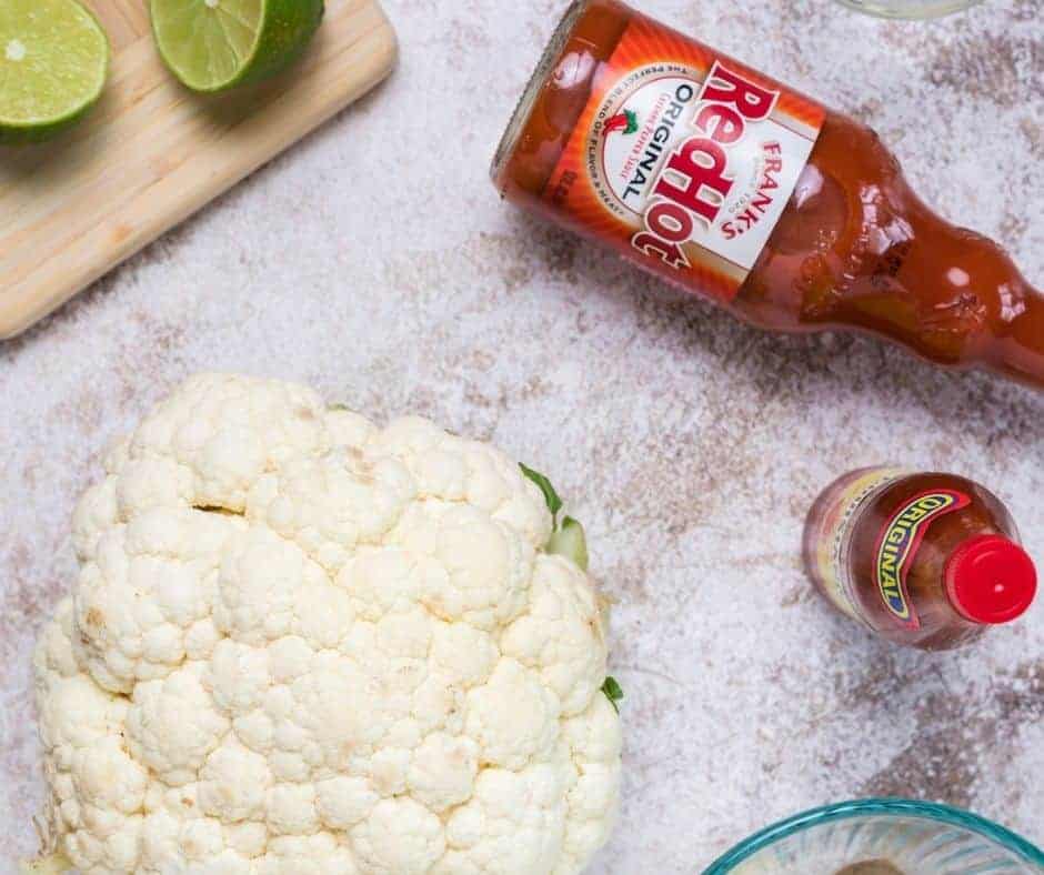 Ingredients Needed For Air Fryer Buffalo Cauliflower Bites sitting on a white marble countertop. 
There is a lime chopped in half on a wooden chipping board. A bottle of Frank's Red Hot Original Sauce. A bottle of Louisiana Hot Sauce and a full cauliflower.