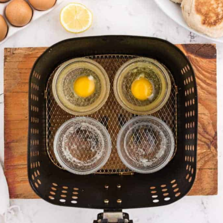 overhead showing how to poach eggs in air fryer: four ramekins in an air fryer basket with two containing raw eggs