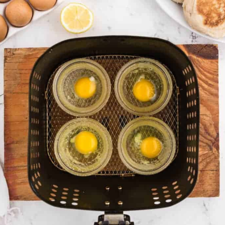 overhead showing how to make poached eggs in air fryer: four ramekins in an air fryer basket with raw eggs in each one