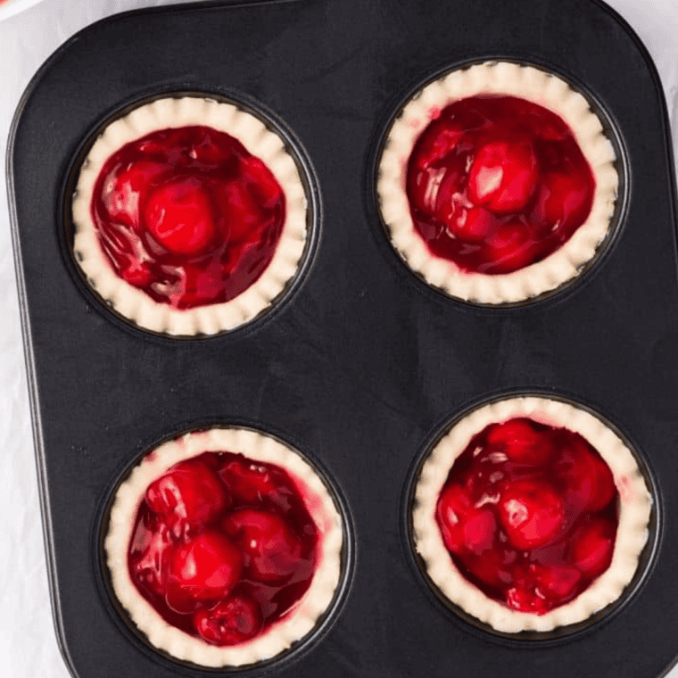 Cherry pie filling being poured into the center of the prepared tart crust in a muffin tin.