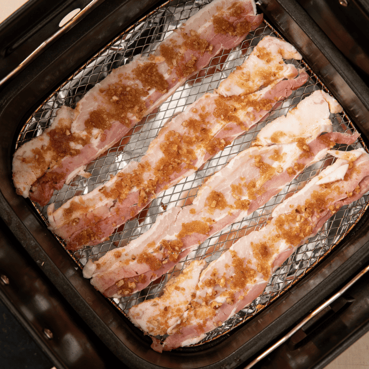 Bacon strips and a sausage patty crisping in an air fryer for a breakfast sandwich.