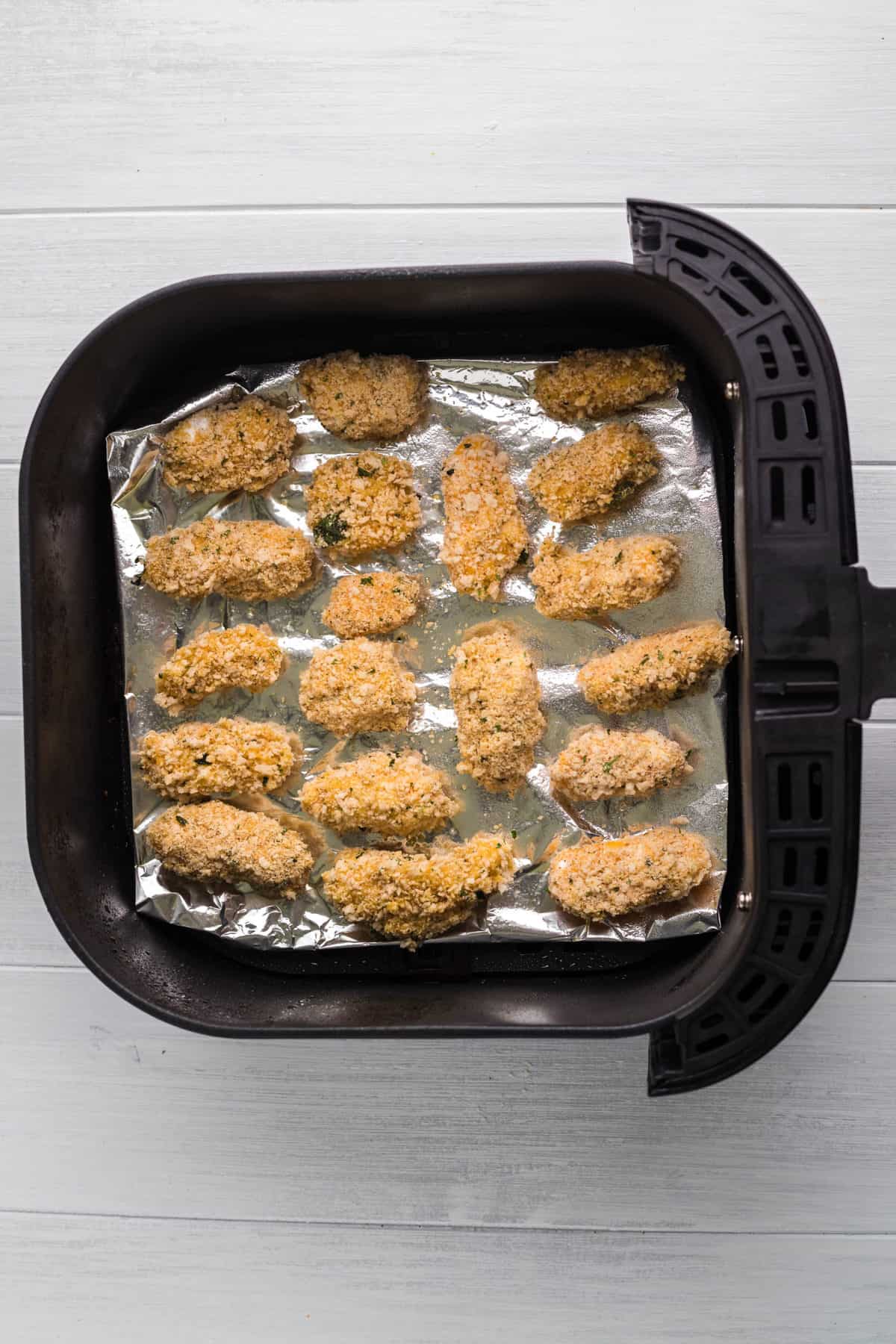 Top view of air fryer basket lined with foil with breaded cheese curds on top of the foil with space around each one. 