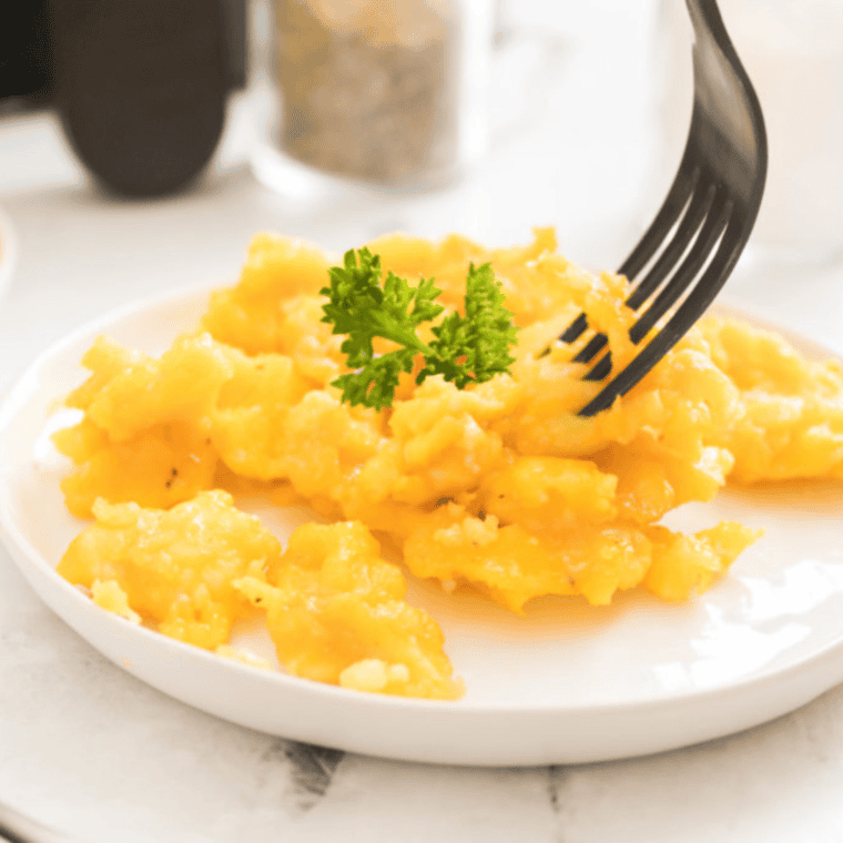 closeup: a fork digging into scrambled eggs made in air fryer on a white plate with fresh parsley on top