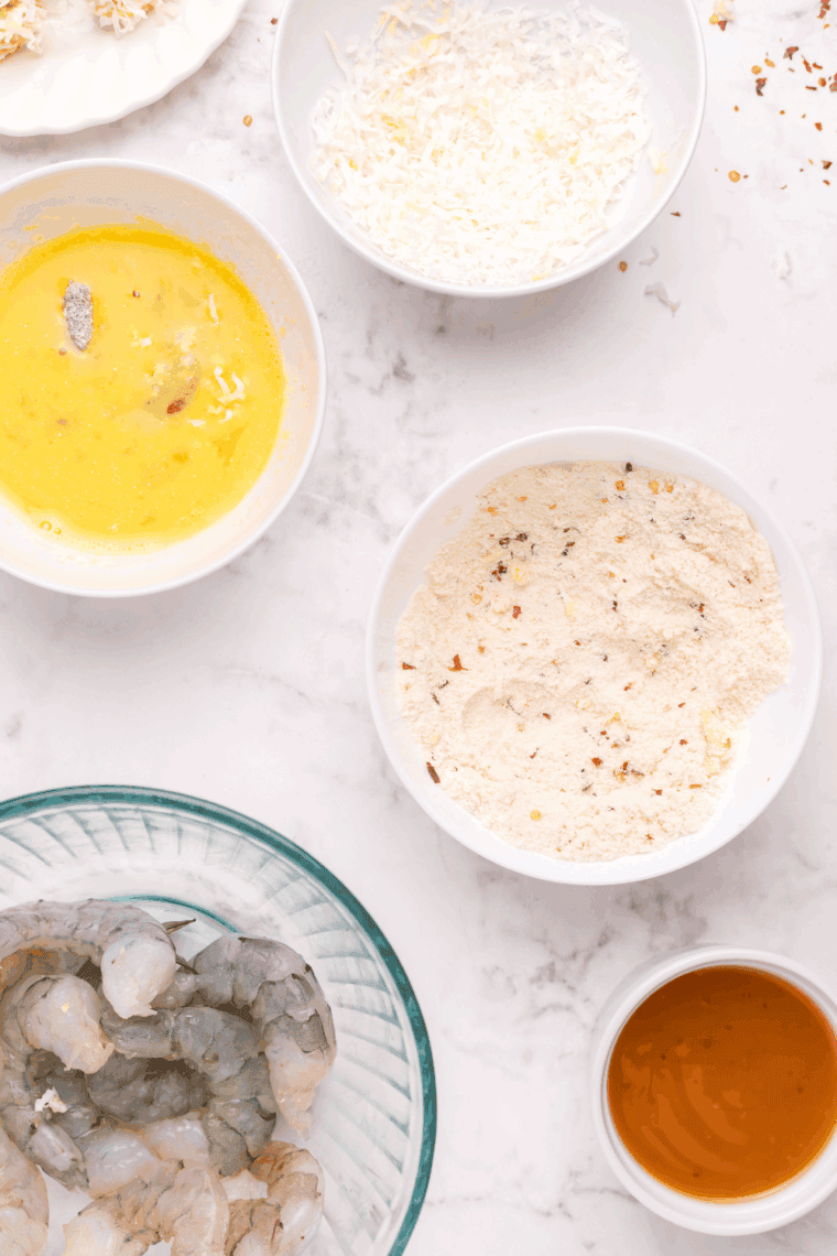 Preparing shrimp by peeling and deveining, with bowls of coconut flour mixture, beaten eggs, and coconut flakes set up for dredging station.