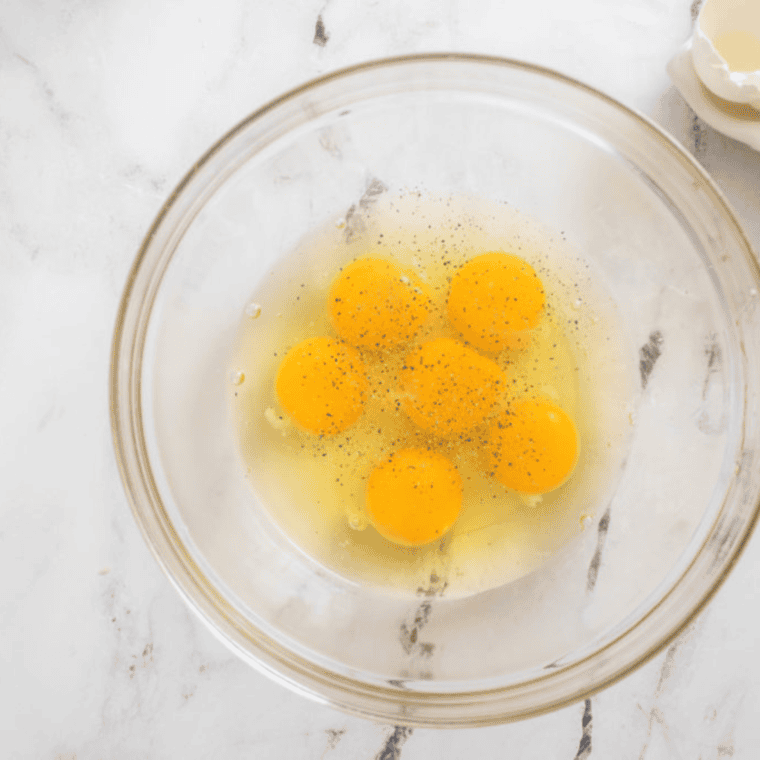 overhead process shot: 6 raw eggs with salt and pepper in a glass bowl before beating them