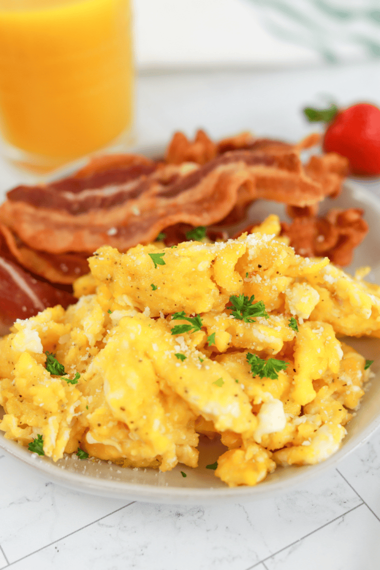 closeup: air fryer scrambled eggs with cheese and fresh herbs on top on a plate with cooked bacon strips